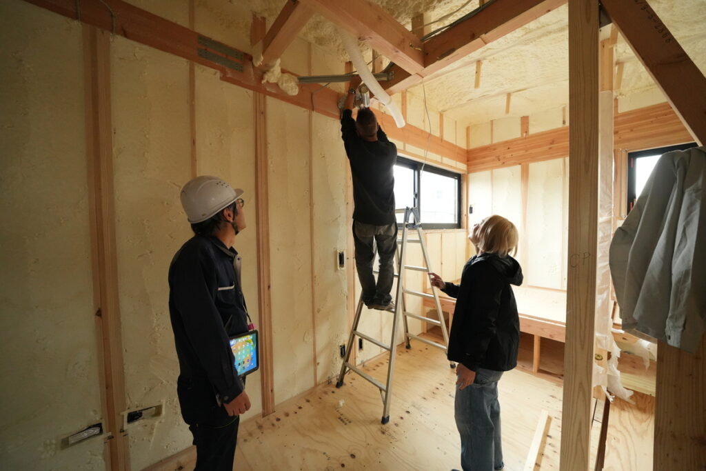 断熱欠損を防ぐ吹き付け断熱の施工現場。専門スタッフが高所作業車や脚立を使用し、天井や壁の細部まで隙間なく施工を確認する。