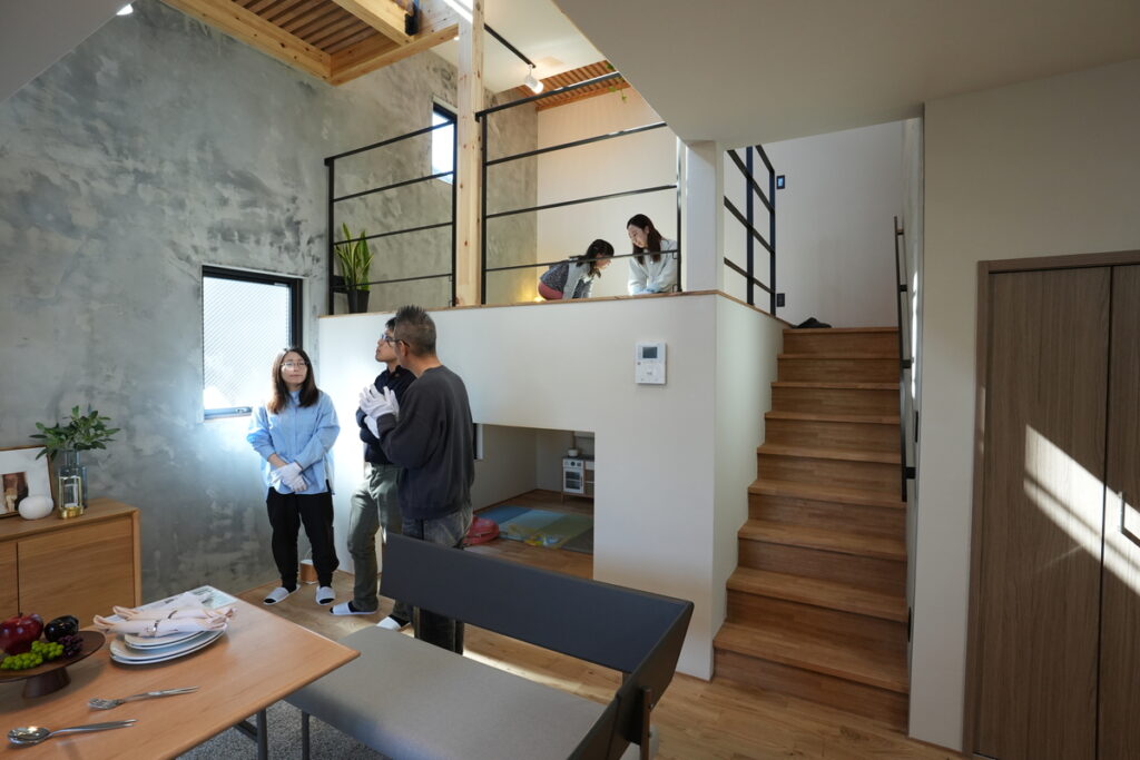 吹き抜けのある開放的なリビングで、夫婦とスタッフが打ち合わせをする様子。モルタル調のアクセントウォールと木目調の天井が調和したモダンな空間。