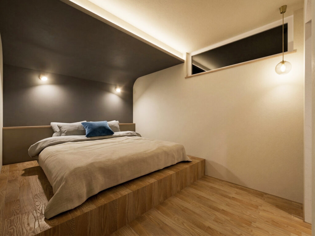 一段高くなったスペースにローベッドを配置した、ホテルライクな主寝室。落ち着いたグレーの塗り壁と、天井の間接照明、ペンダントライトが、心地よい眠りを誘う空間を演出します。