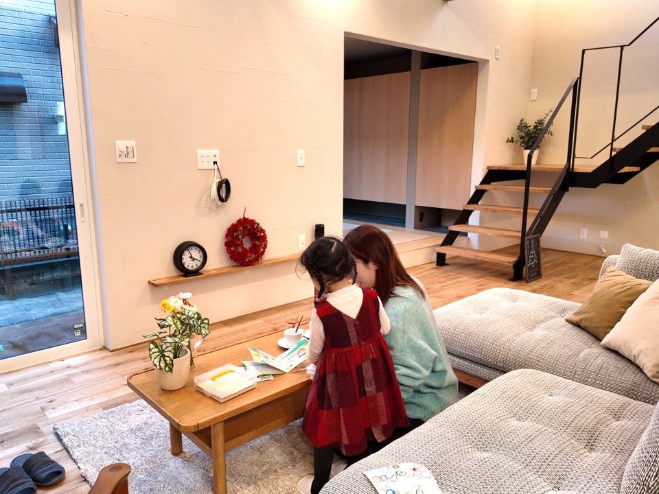 明るい吹き抜けリビングで、スタッフとお客様のお子様がテーブルを囲んで過ごす様子。無垢床やスケルトン階段がある開放的な空間で、お子様連れでも安心して見学や相談ができるアットホームな雰囲気を伝えています。